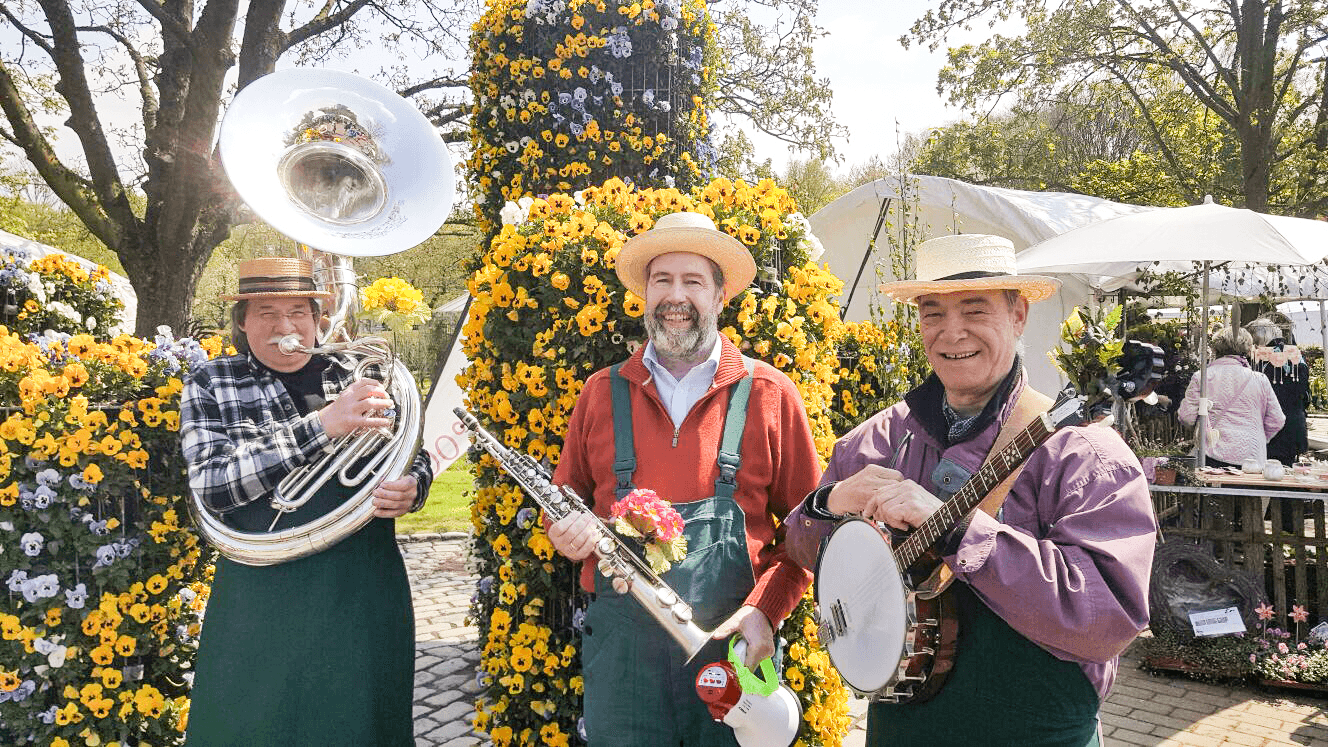 Drei Musiker mit Sousaphon, Sopransaxophon und Banjo vor buntem Blumenstand im Freien.