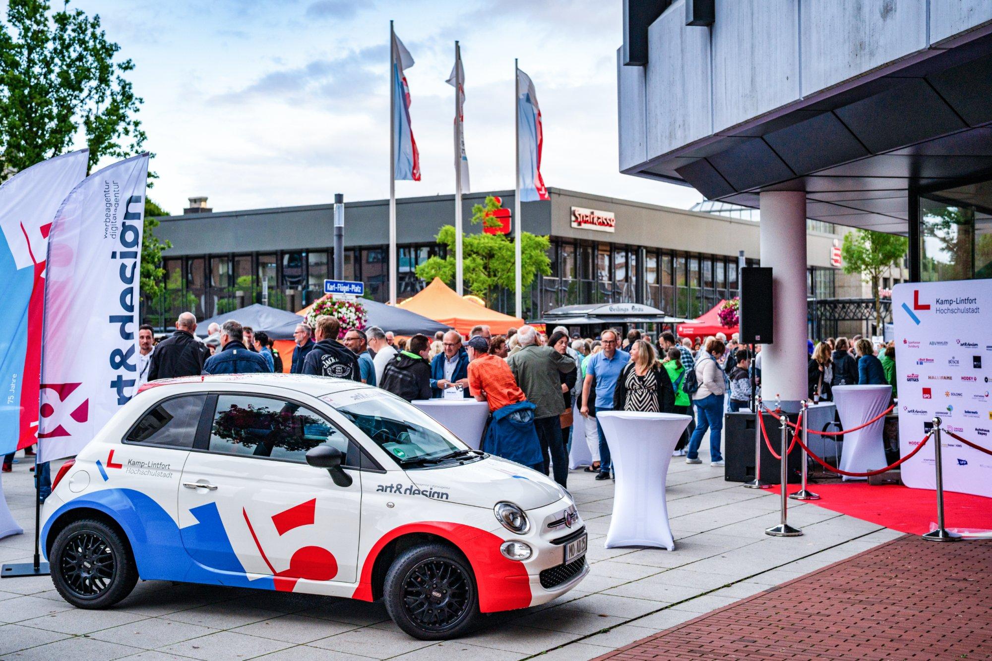 Veranstaltung auf dem Karl-Flügel-Platz in Kamp-Lintfort mit einem weiß-blau-roten Fiat 500 im Stadt Kamp-Lintfort Design im Vordergrund, zahlreichen Besuchern an Stehtischen, Fahnen und Sparkassen-Gebäude im Hintergrund.