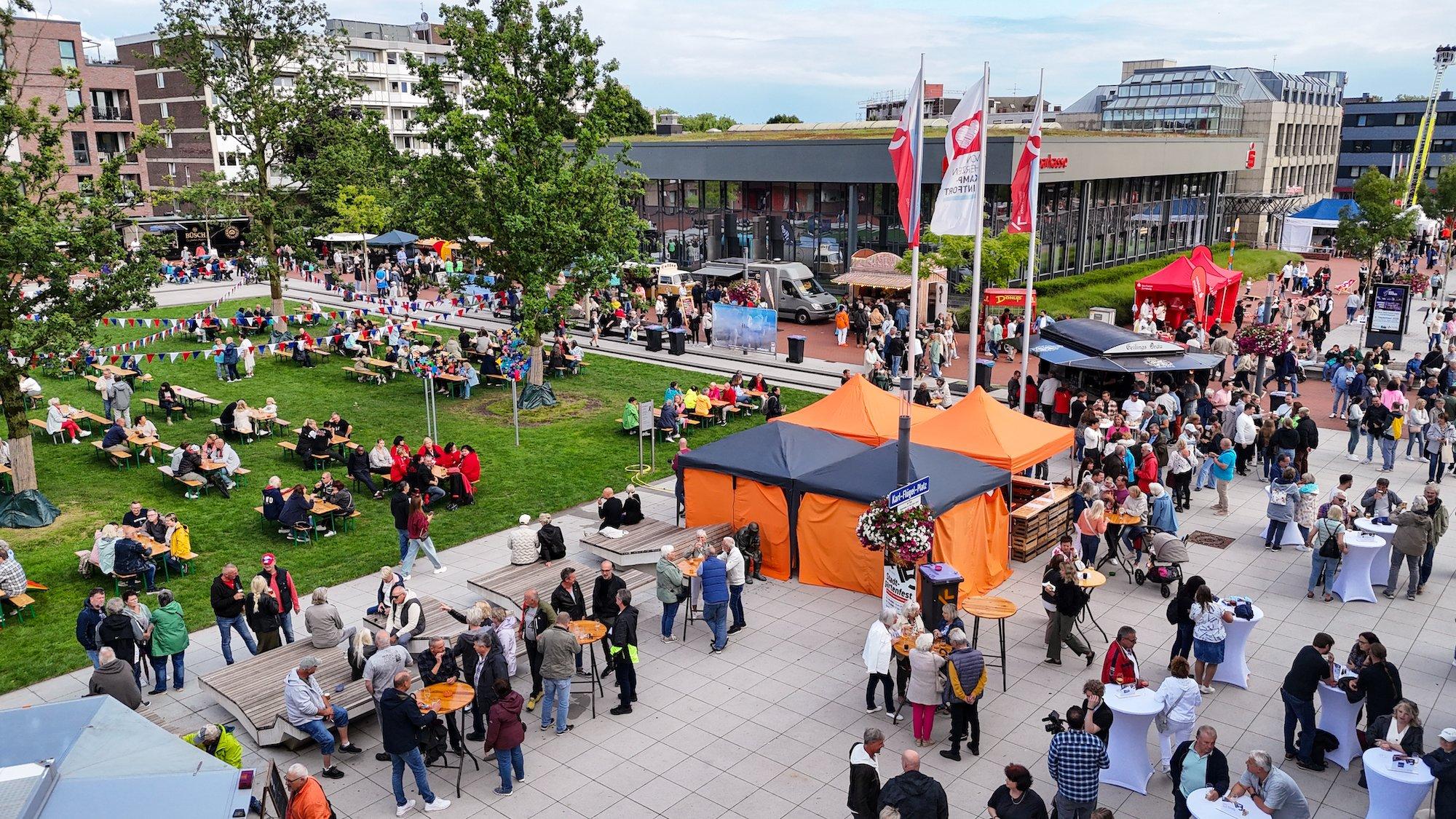 Luftaufnahme eines Stadtfestes auf dem Karl-Flügel-Platz in Kamp-Lintfort mit vielen Besuchern, bunten Pavillons, Bierbänken auf einer Grünfläche, Essensständen und dem Sparkassen-Gebäude im Hintergrund.